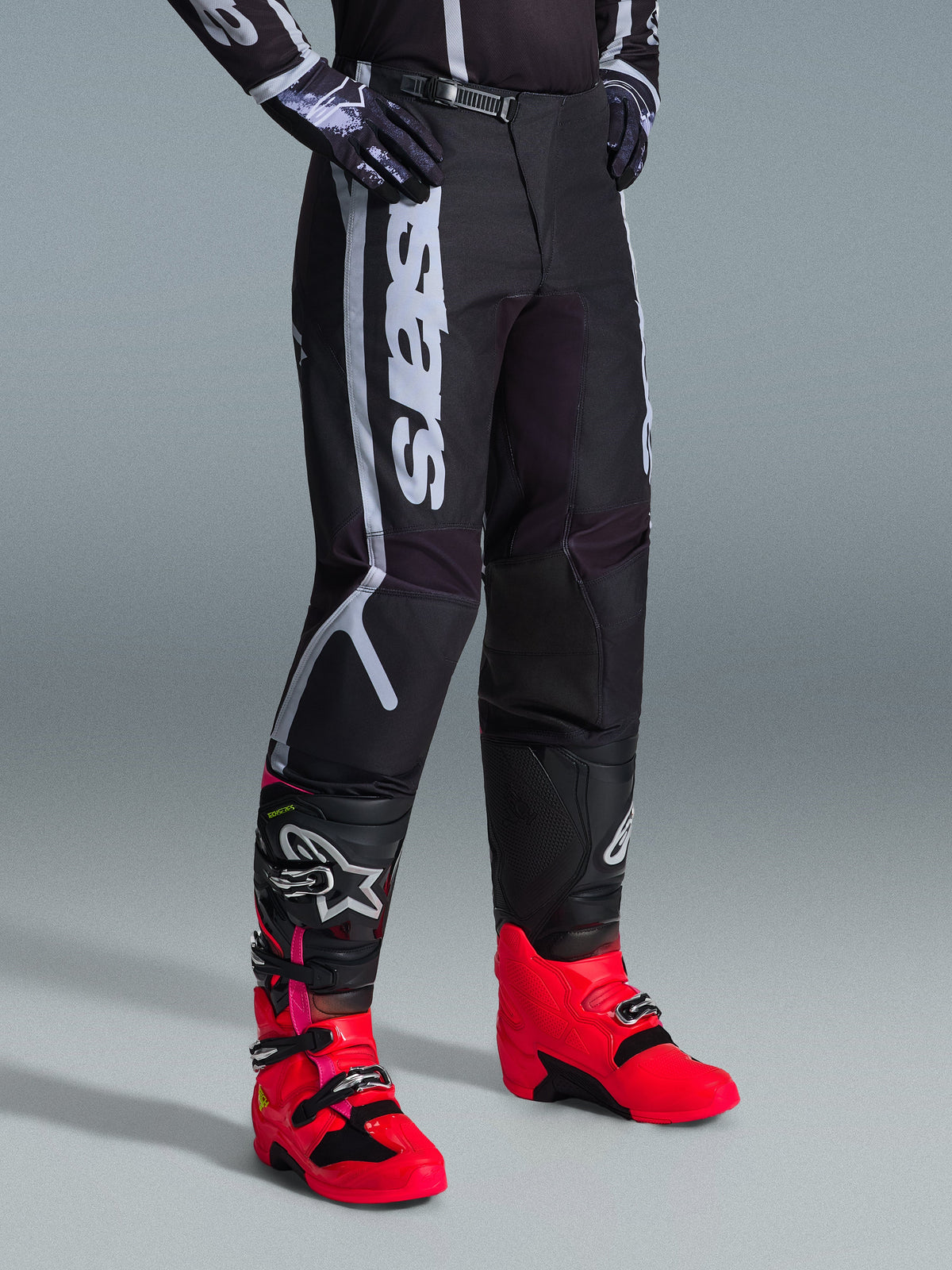 Alpinestars 2026 Fluid Apex Pants, Motocross Pants, Black and Gray, worn with vibrant pink and red boots and matching black and white jersey, featuring bold vertical Alpinestars branding on the thigh, reinforced panels, and ratchet waist closure for off-road performance.