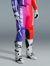 Alpinestars 2026 Fluid Apex Pants, motorcross broek, meerkleurig paars, roze en rood kleurverloop, gedragen met witte en zwarte motorcrosslaarzen en handschoenen, voorzien van verticale witte branding en zwarte knie-accenten, performance off-road uitrusting