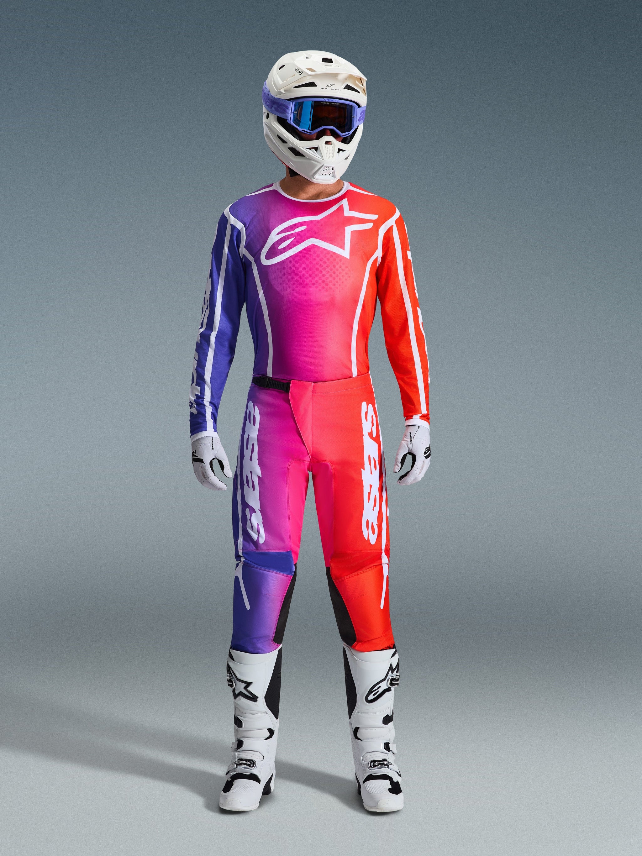 Motorcrosser in 2026 Fluid Apex Pants en bijpassende jersey, paars en oranje kleurverloop, witte helm met bril, witte laarzen en handschoenen, gecoördineerde Alpinestars race-uitrusting voor off-road prestaties.