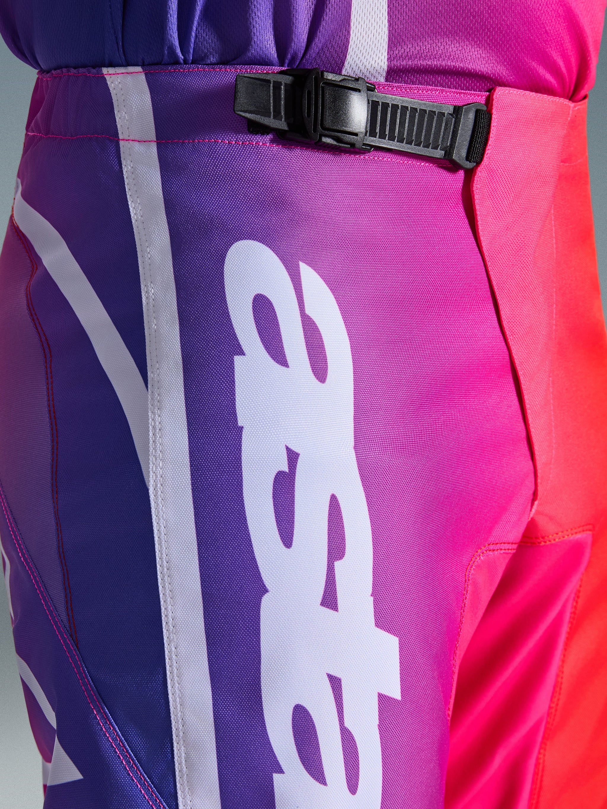 Alpinestars 2026 Fluid Apex Pants, motorcrossbroek, meerkleurig paars, roze en rood, close-up van de taille en heup met een zwarte ratelsluiting en groot wit verticaal Alpinestars-logo, levendig kleurverloop met duurzame polyfabric textuur