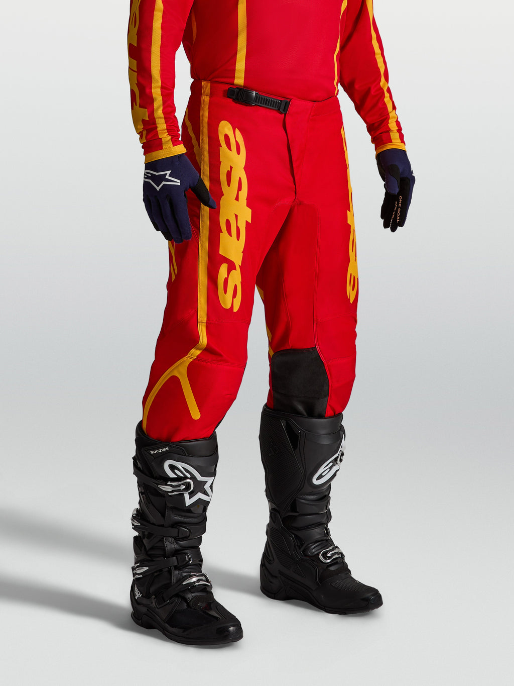 Alpinestars Fluid Apex V2 broek, heren motorcrossbroek in rood en goud, gemaakt van versterkte 600D polystof met ratelsluiting, professionele off-road uitrusting gedragen met zwarte laarzen.