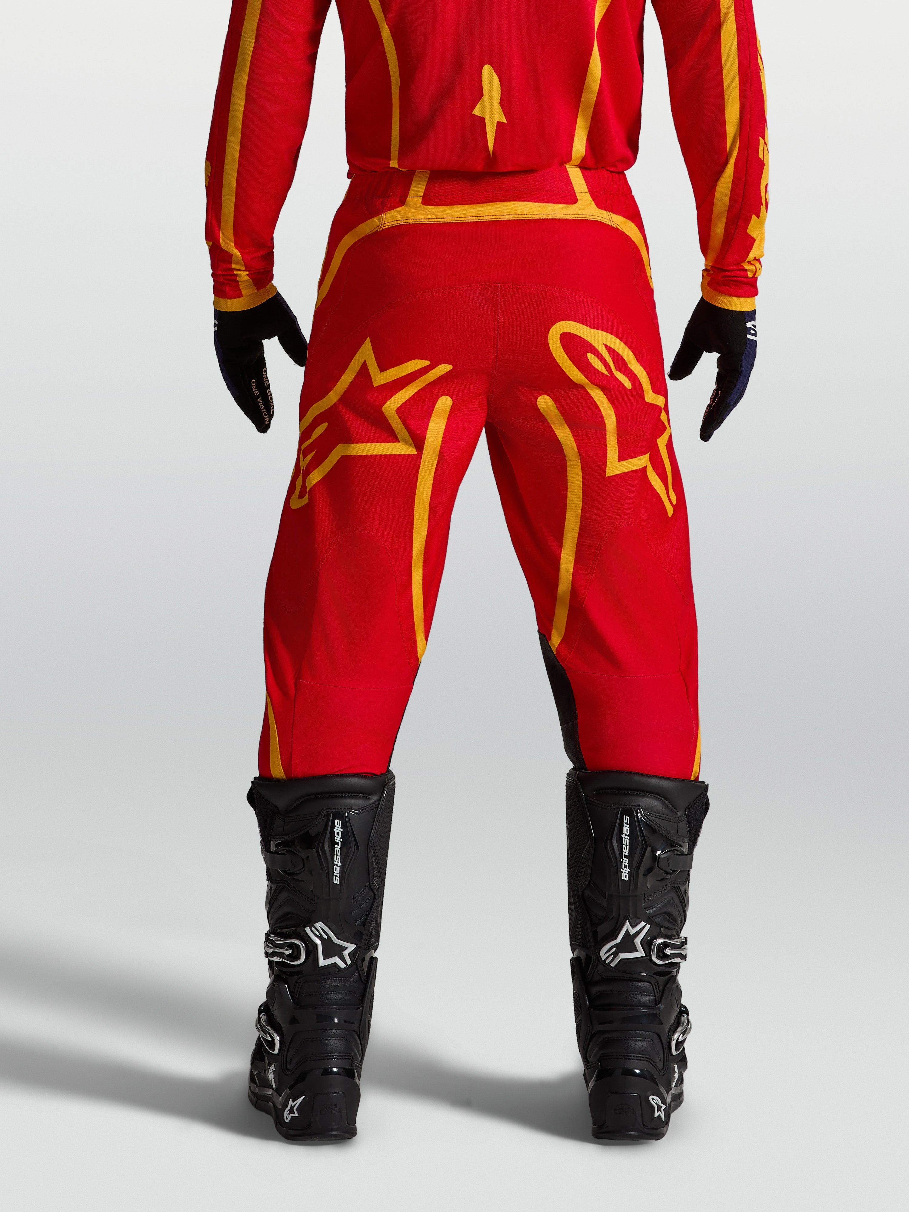 Alpinestars Fluid Apex V2 broek, heren-motocrossbroek in rood en goud, achteraanzicht met zwarte motorlaarzen.