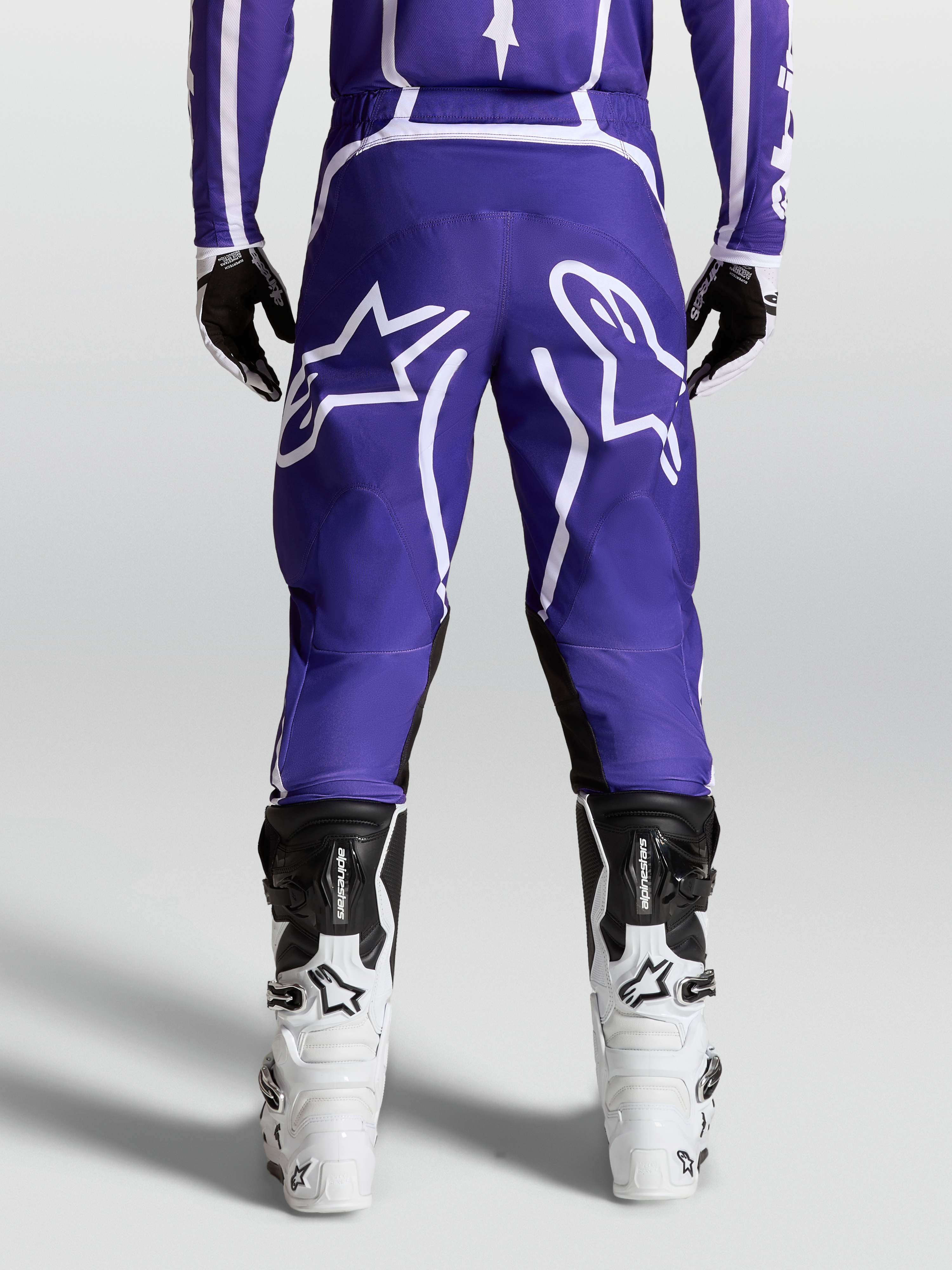 Alpinestars Fluid Apex V2 broek, motocross- en off-roadbroek voor heren, blauw en wit, achteraanzicht op model met motorlaarzen en 600D polyfabric constructie.