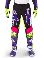 Alpinestars Limited Edition Supertech Dirt Studios motorbroek, motorcrossbroek, wit, paars en neongeel, voorzien van zwarte panelen met neongele en roze accenten, grote paarse sterlogo's, getoond met bijpassende neongele handschoenen en paarse racelaarzen, duurzame off-road racekleding