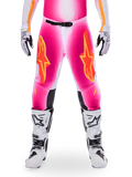 Alpinestars Limited Edition Supertech AFD MIG Pants, motorcrossbroek, fluo roze en wit met oranje accenten, met grote sterlogo's op de bovenbenen, geperforeerde panelen en zwarte knie-gripzones, gedragen met witte en zwarte motorcrosslaarzen