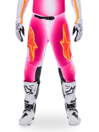 Alpinestars Limited Edition Supertech AFD MIG Pants, motorcrossbroek, fluo roze en wit met oranje accenten, met grote sterlogo's op de bovenbenen, geperforeerde panelen en zwarte knie-gripzones, gedragen met witte en zwarte motorcrosslaarzen