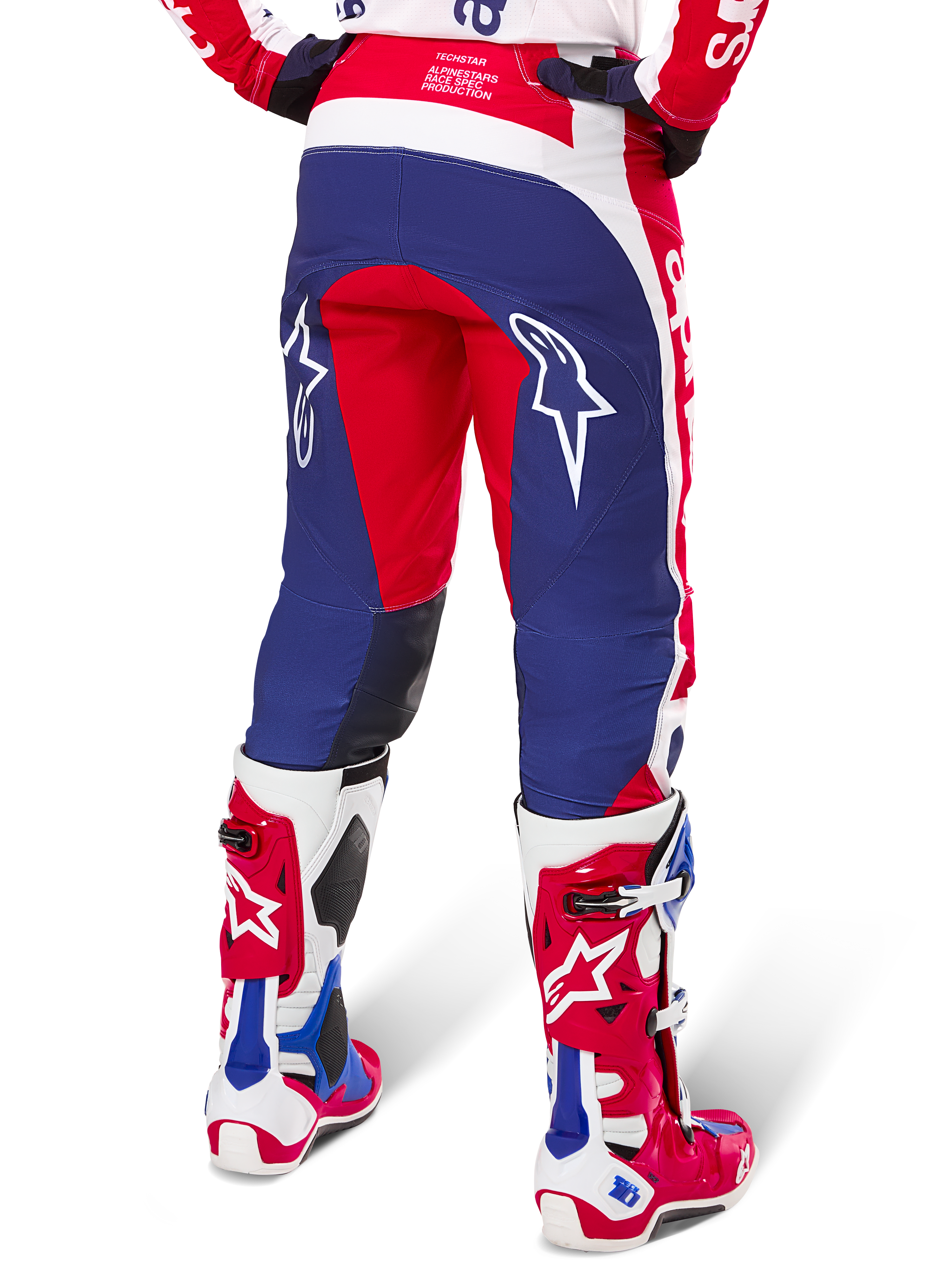 Alpinestars Limited Edition Techstar Fortitude broek, motorcrossbroek, rood, wit en blauw, achteraanzicht gedragen met bijpassende rode en witte laarzen, voorzien van witte sterlogo's op de dijen, duurzaam meerpaneel-ontwerp voor professioneel off-road racen.