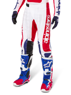 Alpinestars Limited Edition Techstar Fortitude crossbroek, motorcross broek, helder rood, wit en blauw, gedragen met bijpassende off-road laarzen en handschoenen, voorzien van verticale branding op de broekspijp, ademende geperforeerde panelen en zwarte verstevigde kniestukken, atletisch race-ontwerp