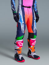 Alpinestars Limited Edition Supertech Records crossbroek, motorcross broek, multicolor met levendig roze en oranje spray-paint effect, marineblauwe en groene panelen, witte logo's, gedragen met bijpassende racelaarzen en handschoenen voor een complete off-road outfit