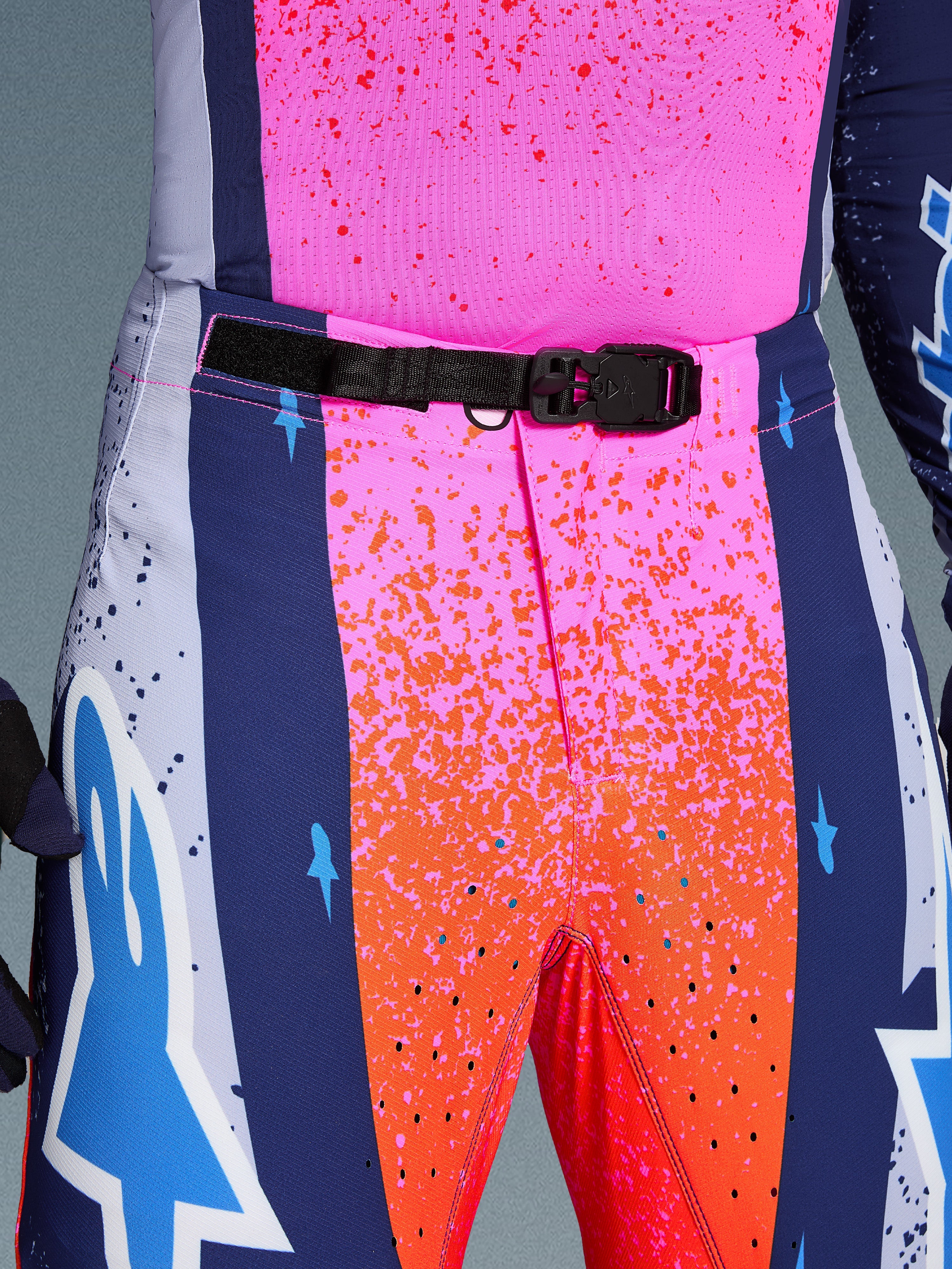 Alpinestars Limited Edition Supertech Records Pants, Motocross Pants, Multicolor pink and orange with navy blue side panels, close-up view of the waist featuring a black SX1 auto-locking magnetic buckle, rip-stop fabric with splatter graphics and laser-cut perforations.