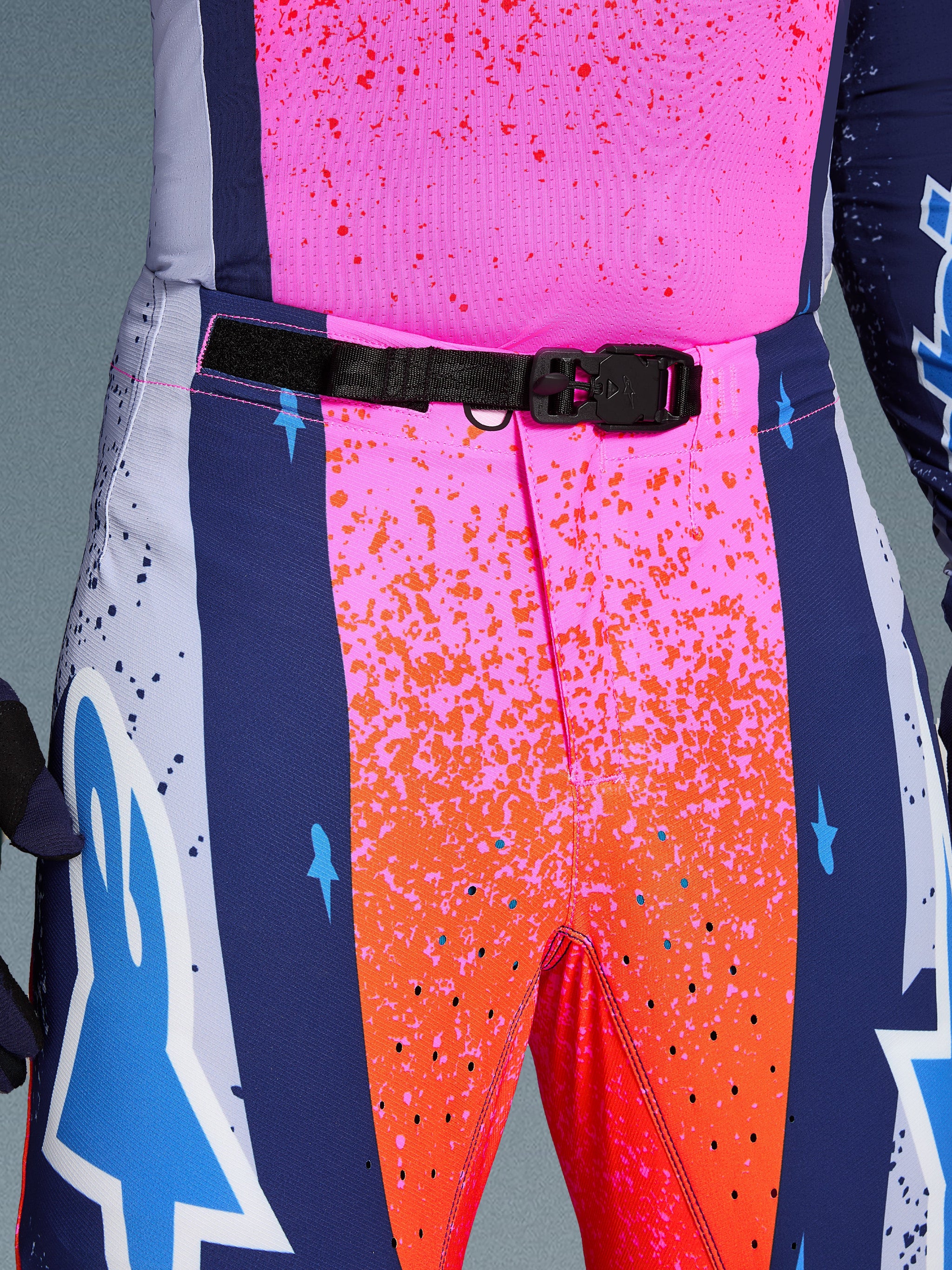 Alpinestars Limited Edition Supertech Records broek, motorcrossbroek, meerkleurig roze, oranje en blauw, close-up van de tailleband en voorsluiting, voorzien van een zwarte verstelbare tailleband met een zelfvergrendelende magnetische gesp, levendig spatpatroon en opvallende graphics voor professionele wedstrijden