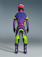 Motocross rider from behind wearing the 2026 Supertech Vista Jersey and matching pants, vibrant yellow and purple design with pink gradients, purple boots with white Alpinestars logos, black gloves, and a red and purple racing helmet, professional-grade racing gear set.
