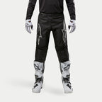 Alpinestars 2024 Fluid Graphite crossbroek, motorcrossbroek, zwart en zilver, met zilveren Alpinestars-logo's op de dijbenen, gedragen met witte en zwarte motorcrosslaarzen en -handschoenen, sportief ontwerp voor off-road racen