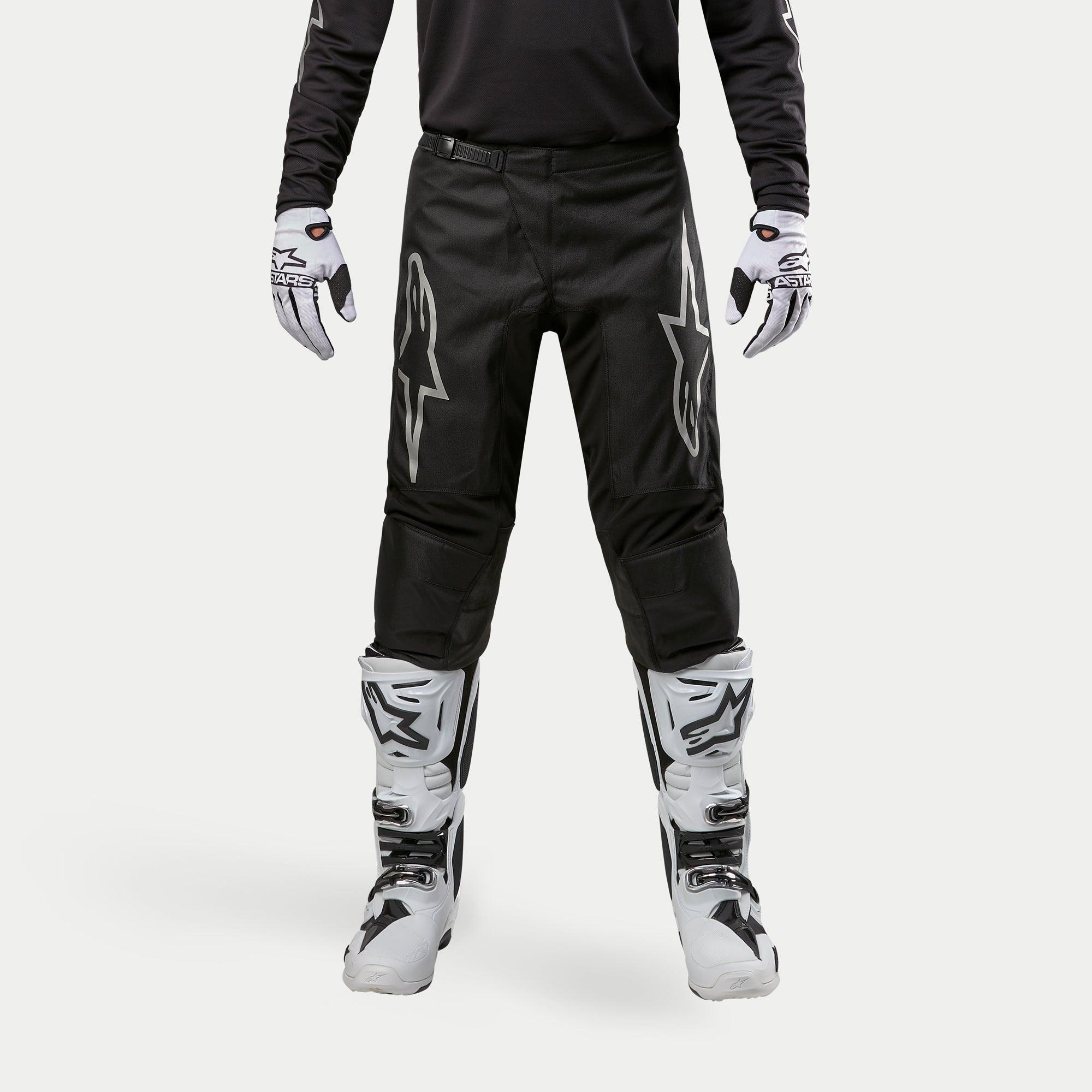 Alpinestars 2024 Fluid Graphite crossbroek, motorcrossbroek, zwart en zilver, met zilveren Alpinestars-logo's op de dijbenen, gedragen met witte en zwarte motorcrosslaarzen en -handschoenen, sportief ontwerp voor off-road racen
