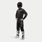 Alpinestars 2024 Fluid Graphite Pants, motorcross broek, zwart en zilver, gedragen met bijpassende zwarte jersey, zwarte helm, witte handschoenen en witte laarzen, voorzien van zilveren Alpinestars-logo's op de dijen, ergonomische sportieve pasvorm voor off-road racen.