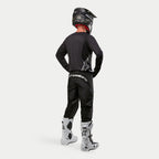 Alpinestars 2024 Fluid Graphite crossbroek, motorcross broek, zwart en zilver, achteraanzicht van een rijder met een zwart shirt, zwarte helm en wit-zwarte laarzen, voorzien van zilveren branding op de onderrug en een duurzame poly-stof constructie voor off-road racing