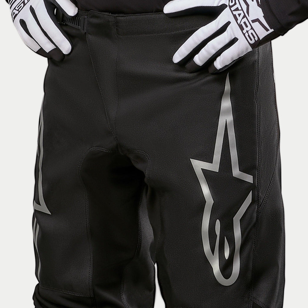 Alpinestars 2024 Fluid Graphite crossbroek, motorcross broek, zwart en zilver, detailopname van de bovenbenen en de taille, met een groot zilveren ster-logo op het been, gedragen met zwart-witte racehandschoenen