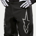 Alpinestars 2024 Fluid Graphite crossbroek, motorcross broek, zwart en zilver, detailopname van de bovenbenen en de taille, met een groot zilveren ster-logo op het been, gedragen met zwart-witte racehandschoenen
