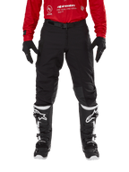 Alpinestars Techdura broek, enduro- en off-roadbroek, zwart, gedragen met een rood Alpinestars shirt en zwart-witte laarzen, voorzien van een robuuste stretch canvas constructie, verstelbare magnetische riemgesp en leren knieverstevigingen voor off-road prestaties.