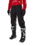 Alpinestars Techdura broek, zwarte off-road motorbroek, gedragen met een rode jersey met lange mouwen en zwart-witte motorcrosslaarzen, robuuste stretch polyamide constructie, leren knieverstevigingen aan de binnenzijde en een verstelbare riem voor enduro rijden.