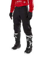 Alpinestars Techdura broek, zwarte off-road motorbroek, gedragen met een rode jersey met lange mouwen en zwart-witte motorcrosslaarzen, robuuste stretch polyamide constructie, leren knieverstevigingen aan de binnenzijde en een verstelbare riem voor enduro rijden.