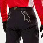 Alpinestars Techdura broek, zwarte endurobroek, achteraanzicht met grote grijze Astars-logo's op het zitvlak, gedragen met een rood shirt en zwarte handschoenen, duurzaam materiaal met zijrits en tailleband met merknaam