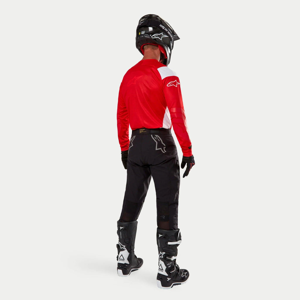 Alpinestars Techdura broek, enduro off-road broek, zwart, achteraanzicht gedragen door een rijder met een rood-wit shirt en zwart-witte laarzen, voorzien van witte sterlogo's op de achterkant en een duurzame technische stretchconstructie.