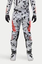 Alpinestars 2024 Techstar Rantera broek, motorcrossbroek, Haze Gray Camo, gedragen met bijpassende jersey, zwarte handschoenen en witte laarzen, voorzien van zwart vlekkenpatroon en rode Alpinestars-branding, duurzaam ontwerp voor off-road racen