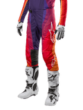 Alpinestars 2024 Techstar Pneuma crossbroek, motorcross broek, paars met oranje en blauw kleurverloop, witte logo's, persoon draagt de broek in combinatie met witte motorcrosslaarzen en handschoenen, sportieve off-road uitrusting