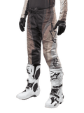 Alpinestars 2024 Techstar Pneuma broek, motorcrossbroek, Dark Sand en Iron Dust grijs, gedragen met witte en zwarte laarzen en zwarte handschoenen, voorzien van een golvend patroon met een groot sterlogo, duurzame uitrusting voor off-road racing