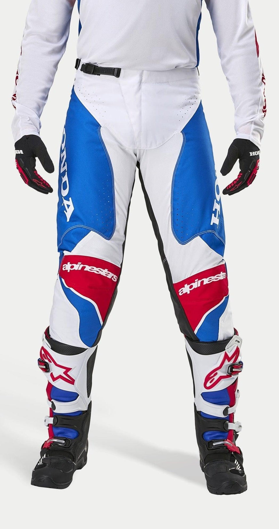 Alpinestars Honda Racer Iconic Pants, motorcross broek, wit, helderblauw en helderrood, met grote Honda-branding op de zijpanelen en het Alpinestars-logo op de knieën, gedragen met bijpassende jersey, zwarte handschoenen en motorcrosslaarzen, ontworpen voor off-road raceprestaties.