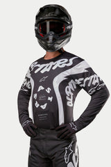 Alpinestars 2024 Racer Hana Jersey, motocross shirt, zwart en wit, voorzien van opvallende witte Alpinestars tekstgraphics en logo, gedragen door een rijder met een zwarte helm en crossbril, hoogwaardige uitrusting voor off-road racing