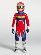 Alpinestars Jeugd Supertech Dyna broek, motocrossuitrusting in rood en marineblauw met witte motorlaarzen en helm.