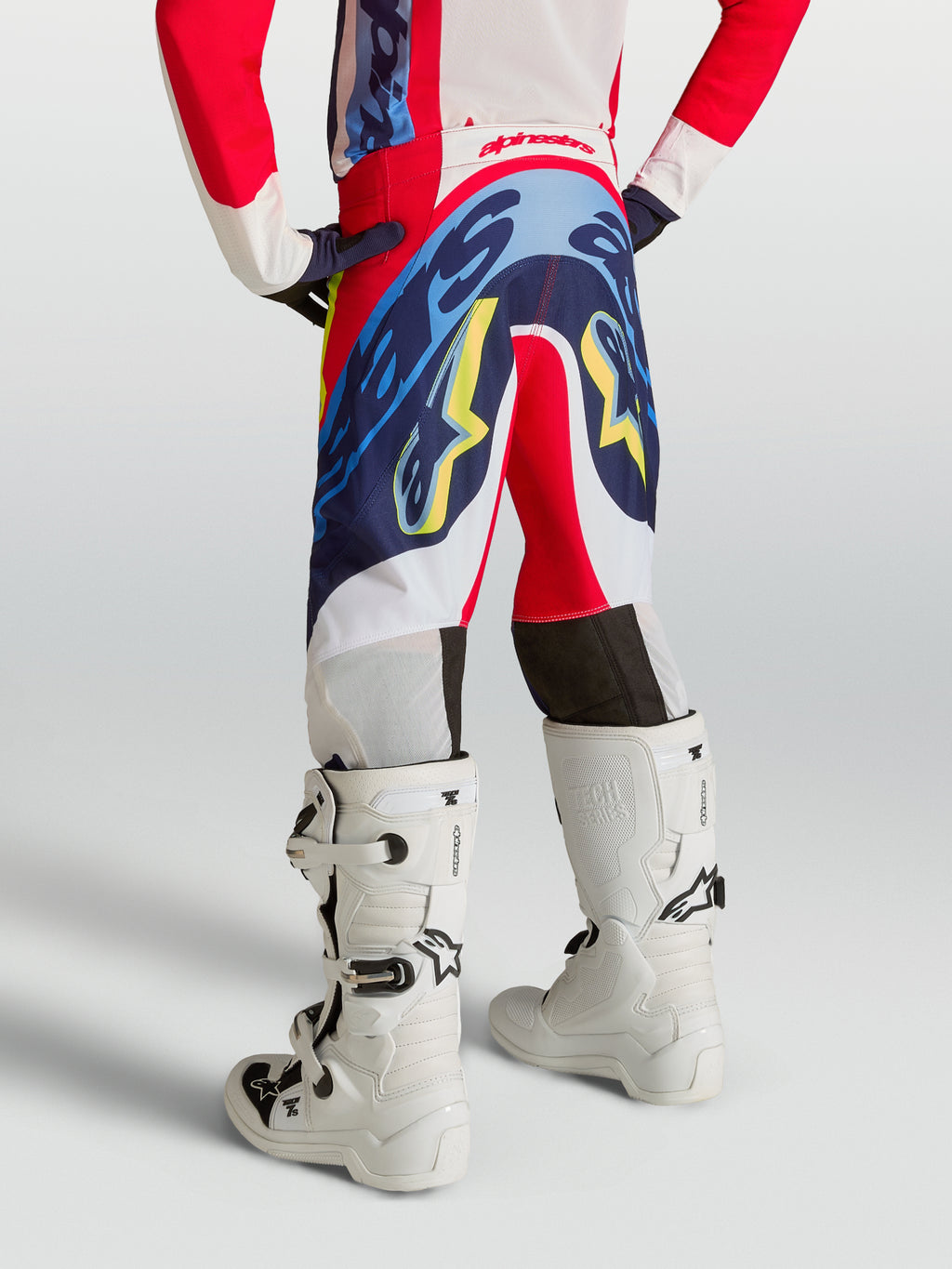 Alpinestars jeugd Supertech Dyna broek, motocrossbroek voor jonge rijders, achteraanzicht, rood, navy blauw en wit, gedragen met witte motorlaarzen, professionele off-road race-uitrusting.