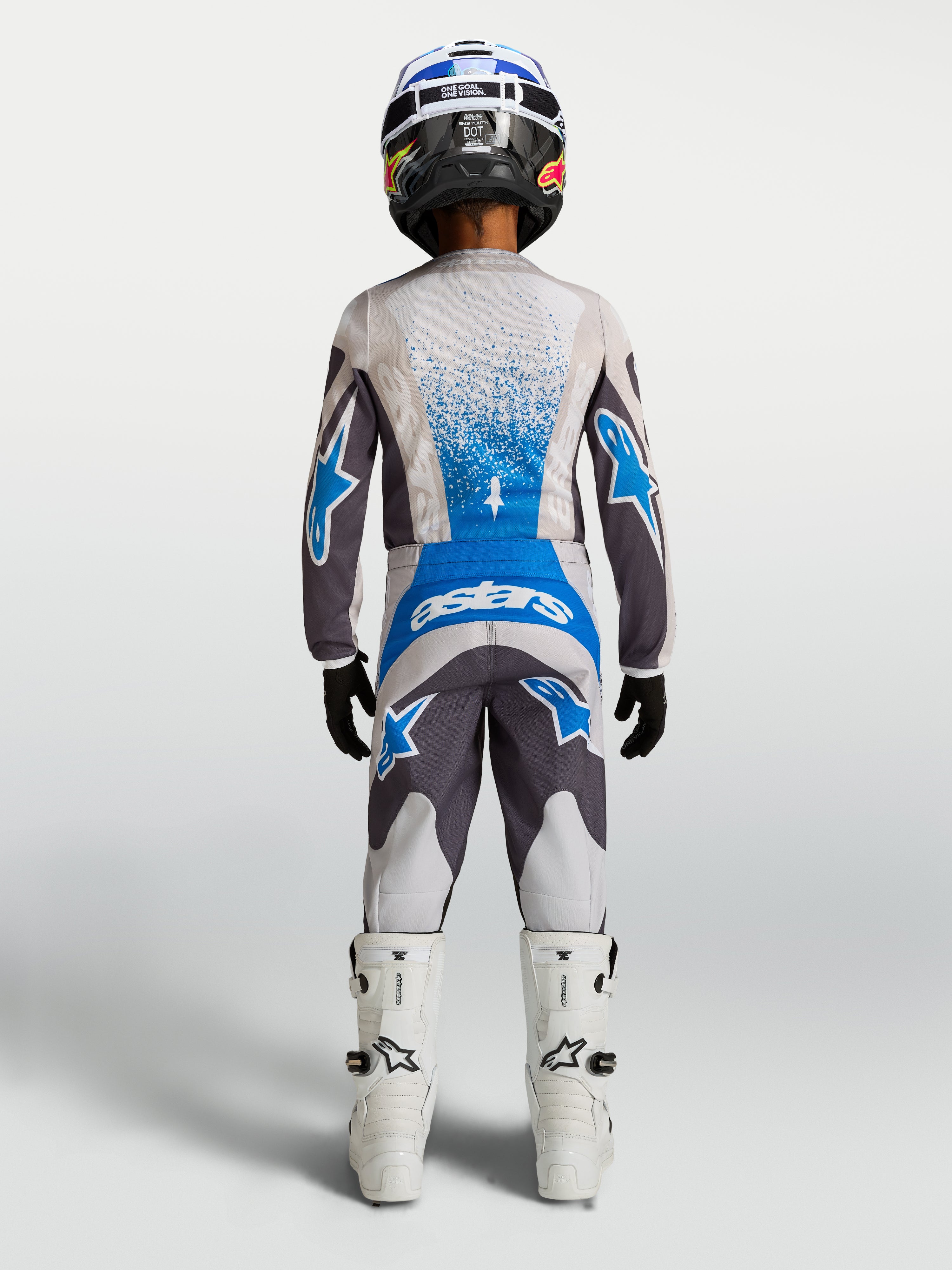 Alpinestars Jeugd Fluid Drag broek, motorcross off-road uitrusting voor jeugd, grijs en blauw, achteraanzicht op model met bijpassende jersey en witte motorlaarzen.