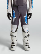 Alpinestars Jeugd Fluid Drag broek 2027, motorcrossbroek, grijs en blauw, 600D polystof met versterkte knieën, gedragen met witte motorlaarzen.