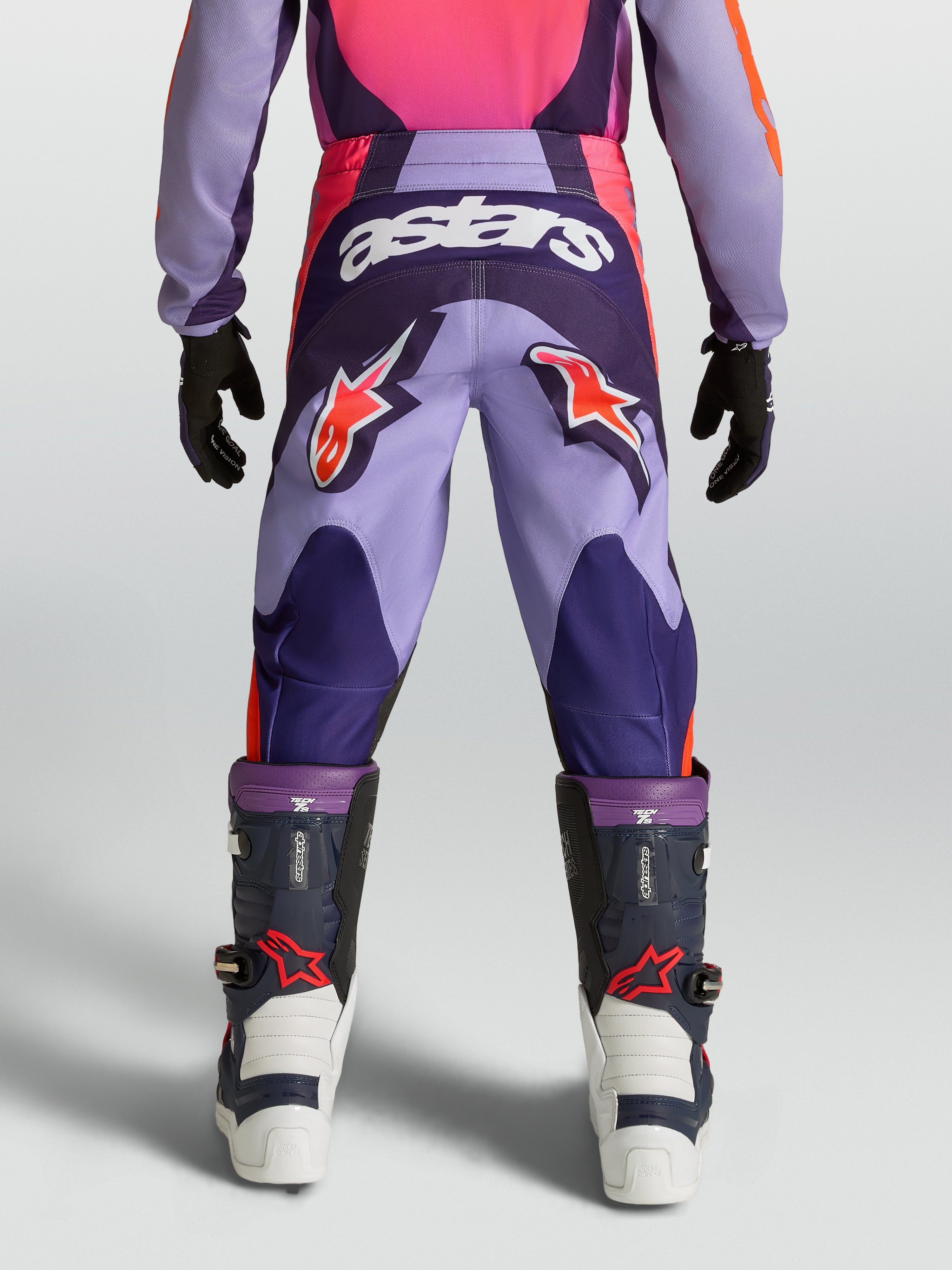 Alpinestars Jeugd Fluid Hunt broek, motocrossbroek voor off-road rijden, paars en roze, duurzame polystof constructie met stretchpanelen, model van achteren gezien met motorlaarzen.