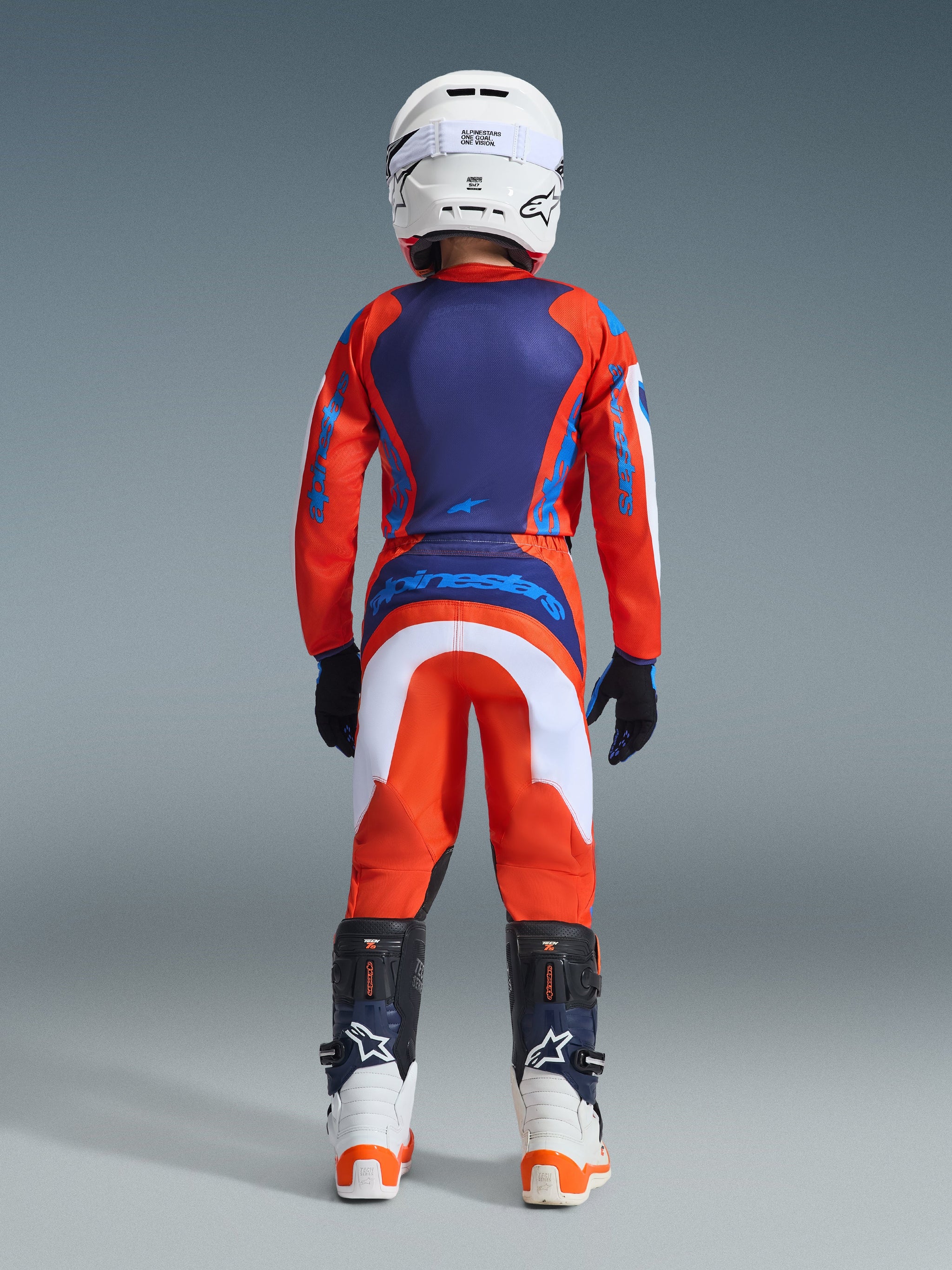 Alpinestars 2026 Youth Fluid Grid Pants, Motocross Pants, Orange and UCLA Blue with white accents, rear view of youth rider in full racing gear including matching jersey, white helmet, and white/blue motocross boots