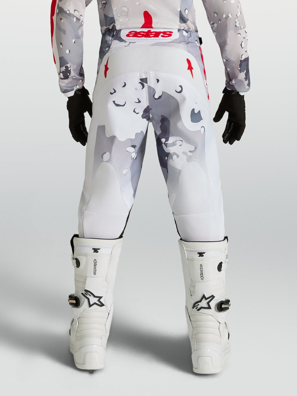 Alpinestars Jeugd Fluid Lone broek, motorcrossbroek voor jeugd, grijs camouflage en wit met rode accenten, achteraanzicht op rijder met witte motorlaarzen.