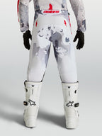 Alpinestars Jeugd Fluid Lone broek, motorcrossbroek voor jeugd, grijs camouflage en wit met rode accenten, achteraanzicht op rijder met witte motorlaarzen.