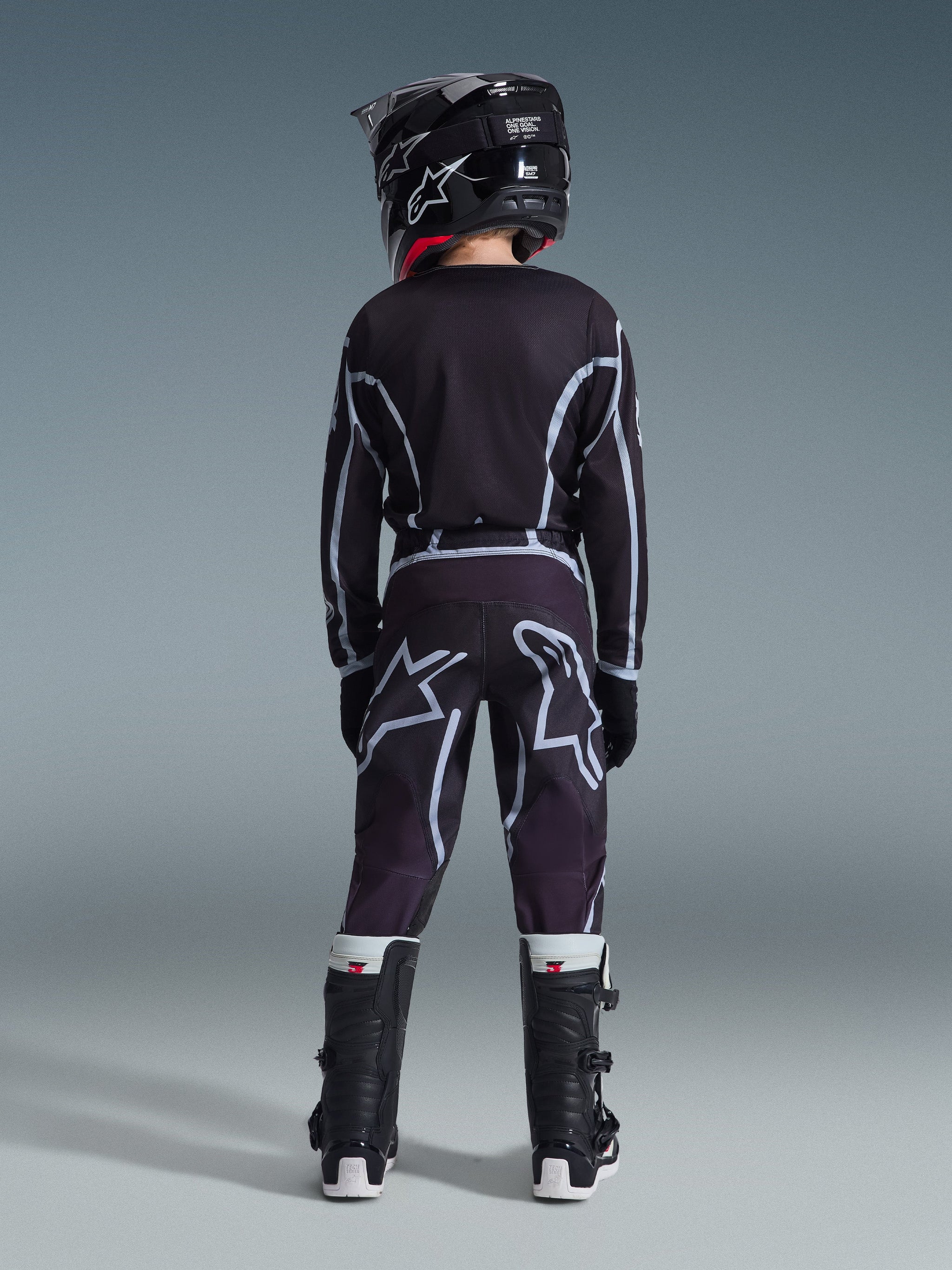 Rear view of a youth motocross rider wearing Alpinestars 2026 Youth Fluid Apex Pants in Black and Gray, matching long-sleeve jersey, black helmet, and black and white motocross boots, coordinated off-road racing gear.