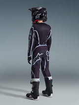Alpinestars 2026 Youth Fluid Apex Pants, Youth Motocross Pants, Black and Gray, worn by a young rider from behind with coordinating black jersey and helmet, featuring bold white logos and geometric patterns, paired with black and white boots for off-road racing.