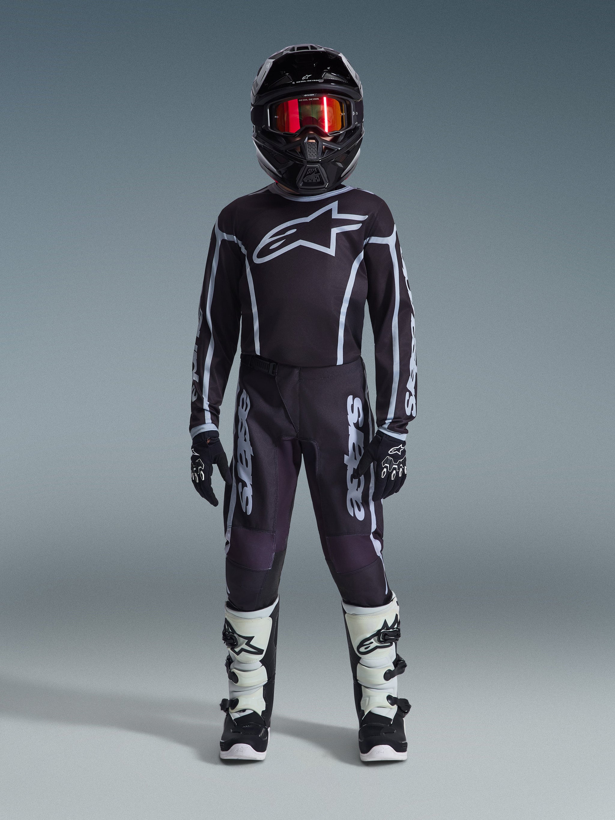 Youth motocross rider wearing complete racing gear, 2026 Youth Fluid Apex Pants in Black and Gray, matching black jersey with white Alpinestars logo, black helmet with red mirrored goggles, white and black boots, black and white gloves, coordinated outfit for off-road racing