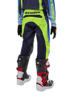 Alpinestars Youth 2024 Racer Hoen motorcrossbroek, fluogeel en donkerblauw, achteraanzicht met opvallende Alpinestars-branding op de tailleband, gedragen met zwart-rode Tech Series laarzen en bijpassende jersey, duurzame sportieve pasvorm voor off-road rijden