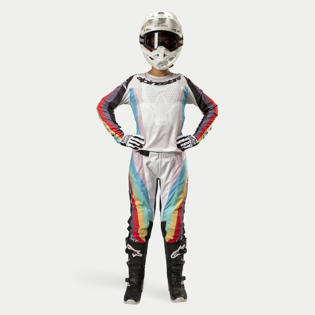Vrouwelijke motorcrosser in Alpinestars Stella 2024 Techstar broek, Black Multicolor, witte basis met rode, gele en blauwe strepen, bijpassende jersey met lange mouwen, witte helm en zwarte laarzen, volledige race-uitrusting voor off-road competitie