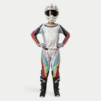 Vrouwelijke motorcrosser in Alpinestars Stella 2024 Techstar broek, Black Multicolor, witte basis met rode, gele en blauwe strepen, bijpassende jersey met lange mouwen, witte helm en zwarte laarzen, volledige race-uitrusting voor off-road competitie