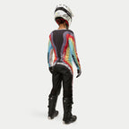 Motorcrosser van achteren gezien in Alpinestars Stella 2024 Techstar damesbroek in Black Multicolor, voorzien van een groot wit logo op het zitvlak, gecombineerd met een kleurrijke gestreepte jersey, witte helm en zwarte Tech 3-laarzen voor off-road racing.