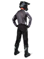 Motocross rider wearing Alpinestars Pro-Dura Jersey in Black Magnet White, rear view showing grey mesh back panel and black sleeves with white Alpinestars branding, coordinated with black pants, black helmet, and black and white boots, professional enduro racing gear