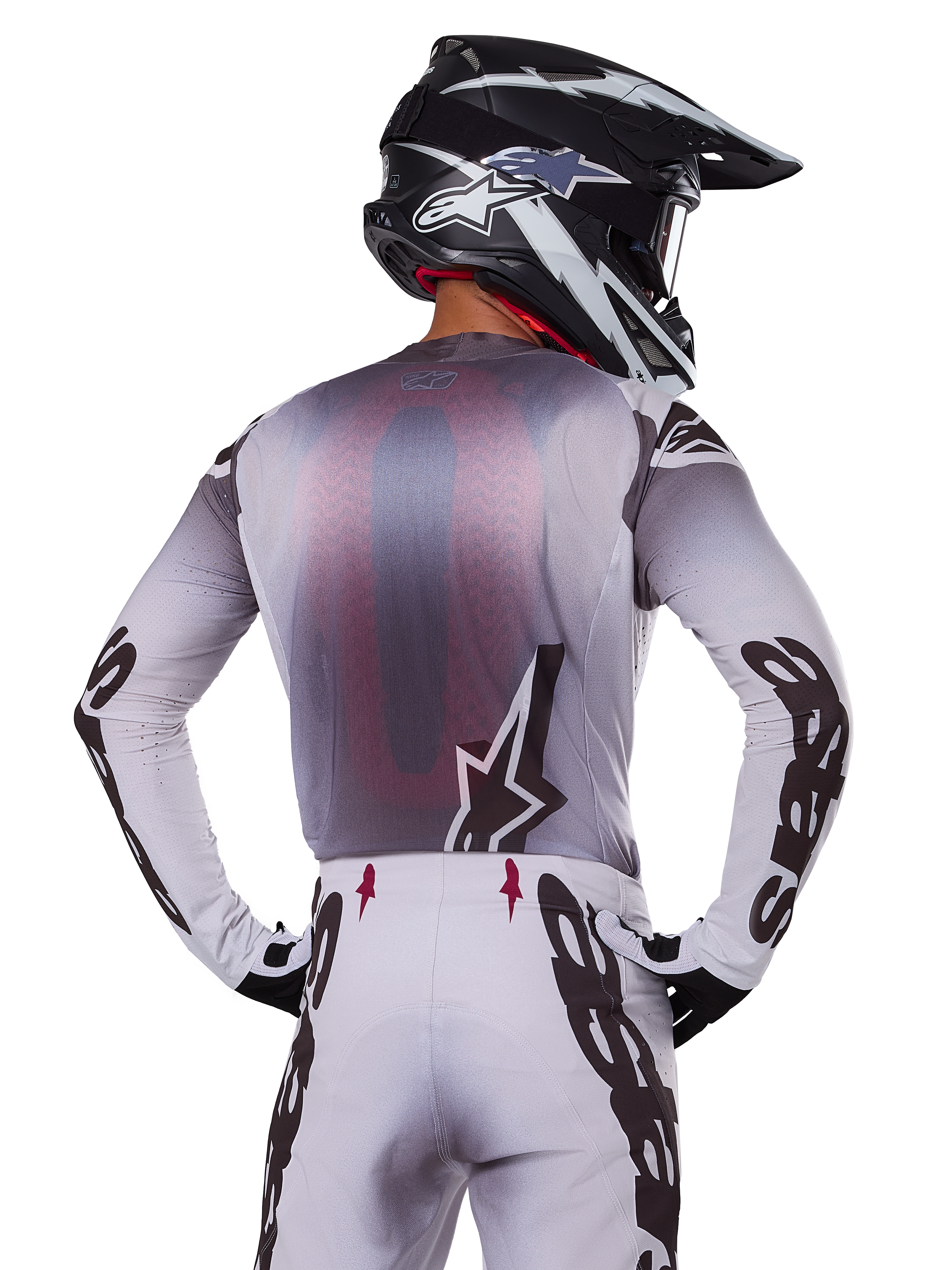 Alpinestars 2025 Supertech Maker Gear Set, motorcrosser van achteren gezien, wit en grijs shirt met grote zwarte merknaam op de mouwen en ster-logo, bijpassende witte broek met zwarte graphics, zwart-witte helm, professionele off-road race-outfit