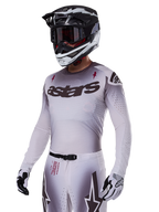 Motorcrosser die de Alpinestars 2025 Supertech Maker Gear Set in het wit draagt, met een jersey met grijs kleurverloop, het zwarte astars-logo en lasergesneden perforaties, bijbehorende broek met magnetische gesp en ster-graphics, gedragen met een zwart-witte helm en handschoenen voor een complete off-road race-look.